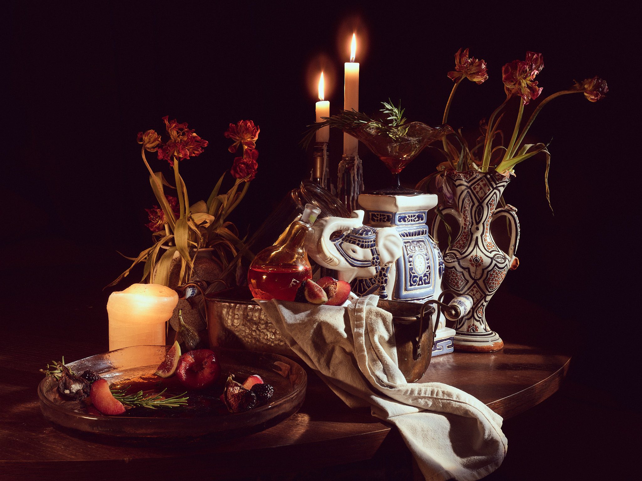 A still life by candlelight, dried roses and tulips going over in stoneware jugs, fruit and a folded cloth on dark wood.