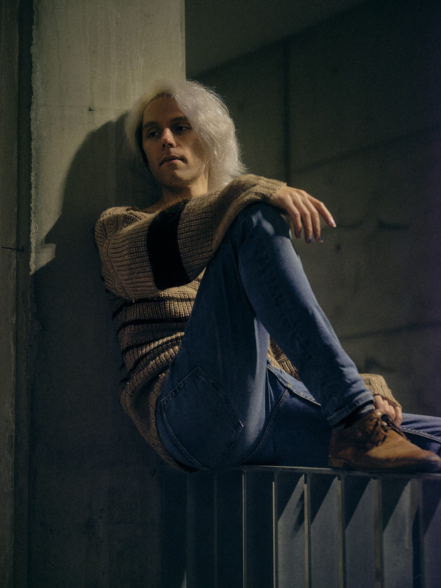 A person with long pale hair sits on a metal railing against a concrete wall, knees drawn up, looking past the camera in cool desaturated light.