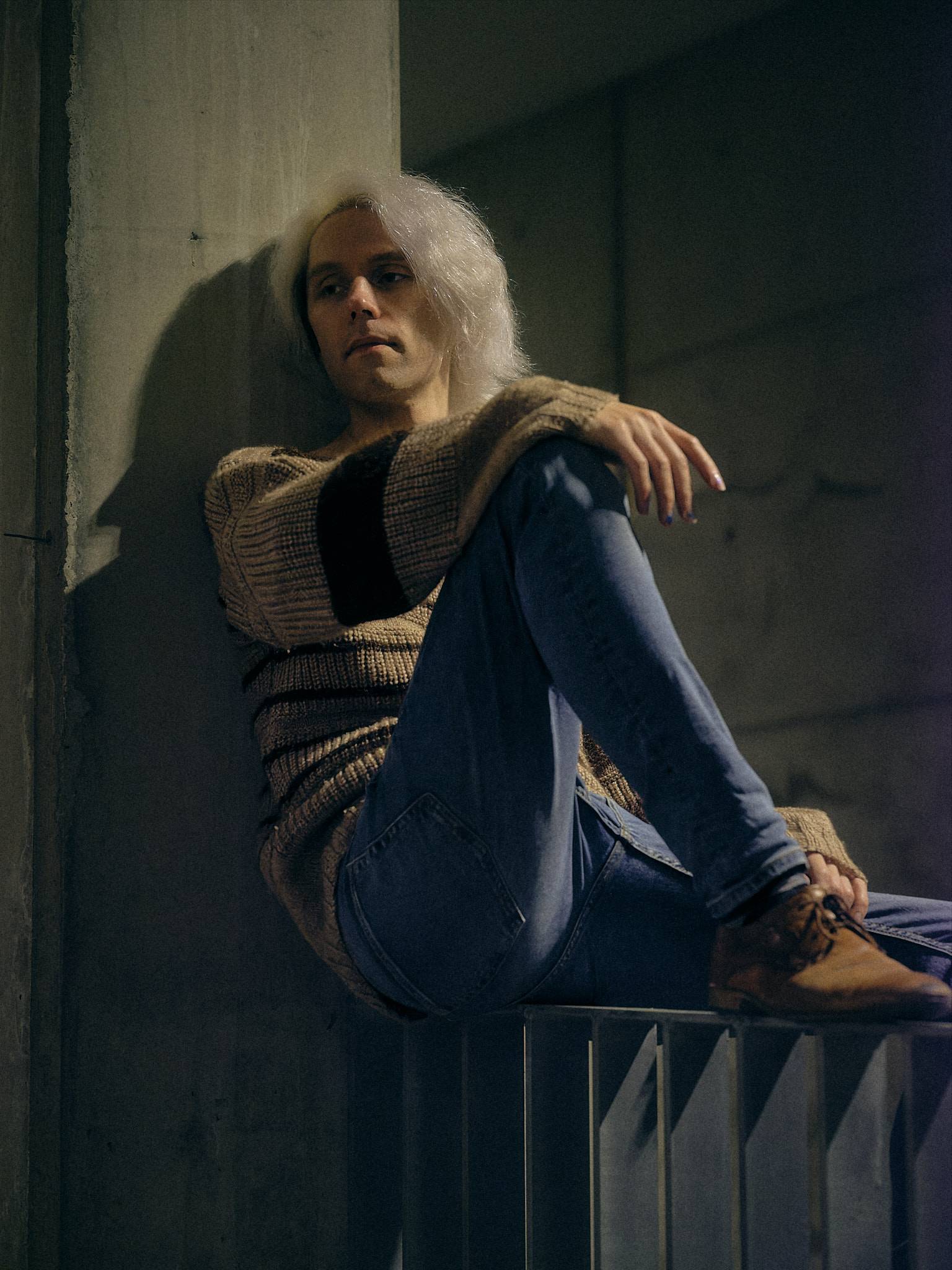 A person with long pale hair sits on a metal railing against a concrete wall, knees drawn up, looking past the camera in cool desaturated light.