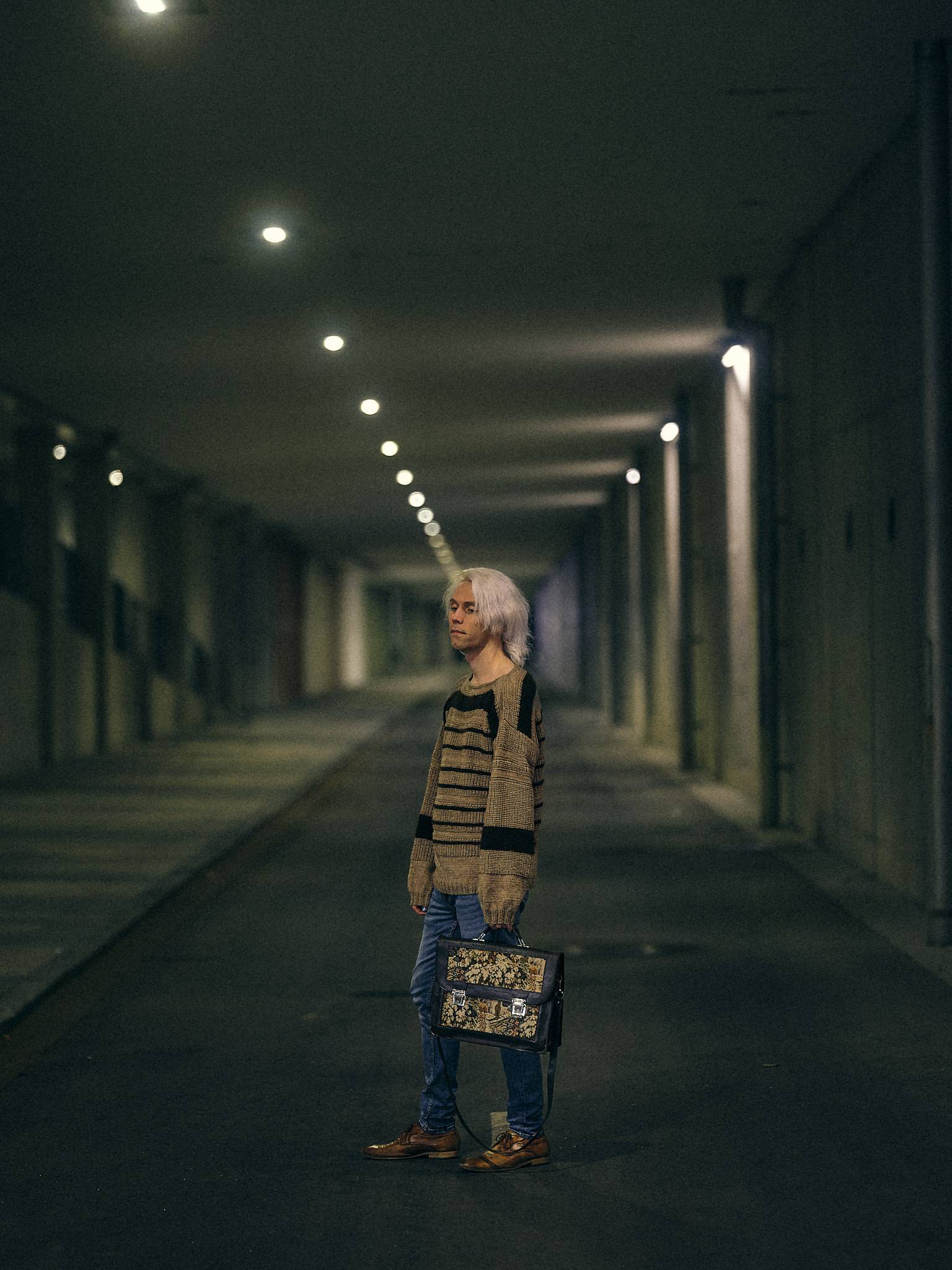 A figure stands alone in a long concrete underpass at night holding a floral tapestry briefcase, a receding line of ceiling lights behind them.