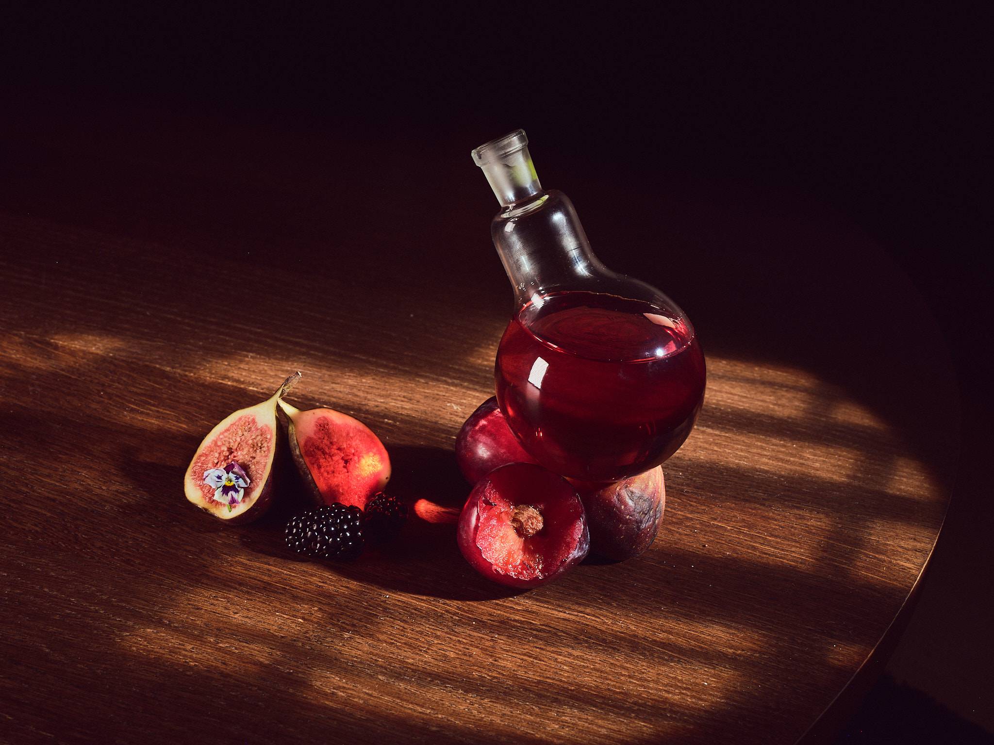 A glass decanter of red wine on a wooden table in low raking light, with a halved fig, blackberries and pomegranates.