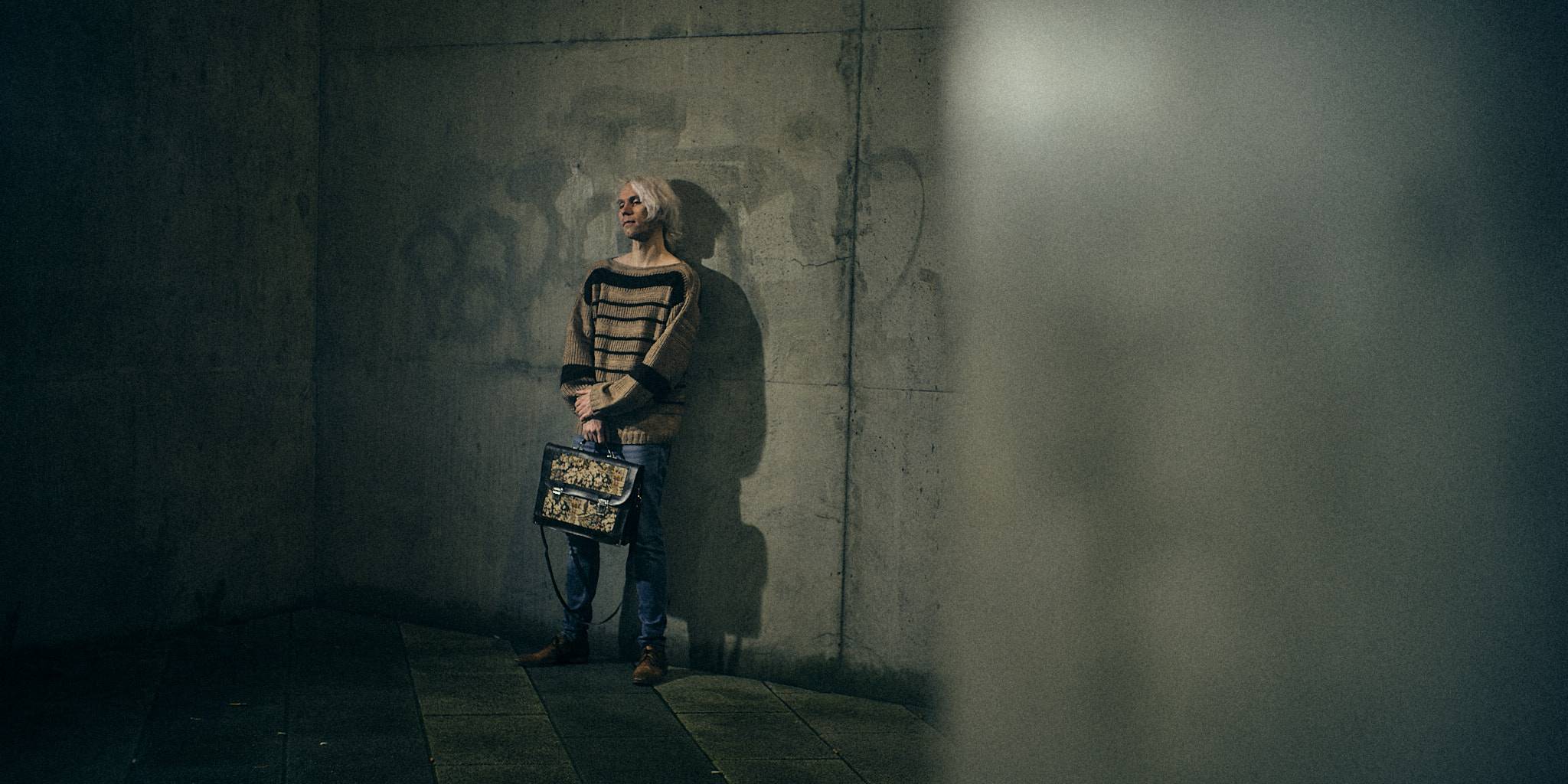 A figure standing far off against a bare concrete wall, small in a wide empty frame.