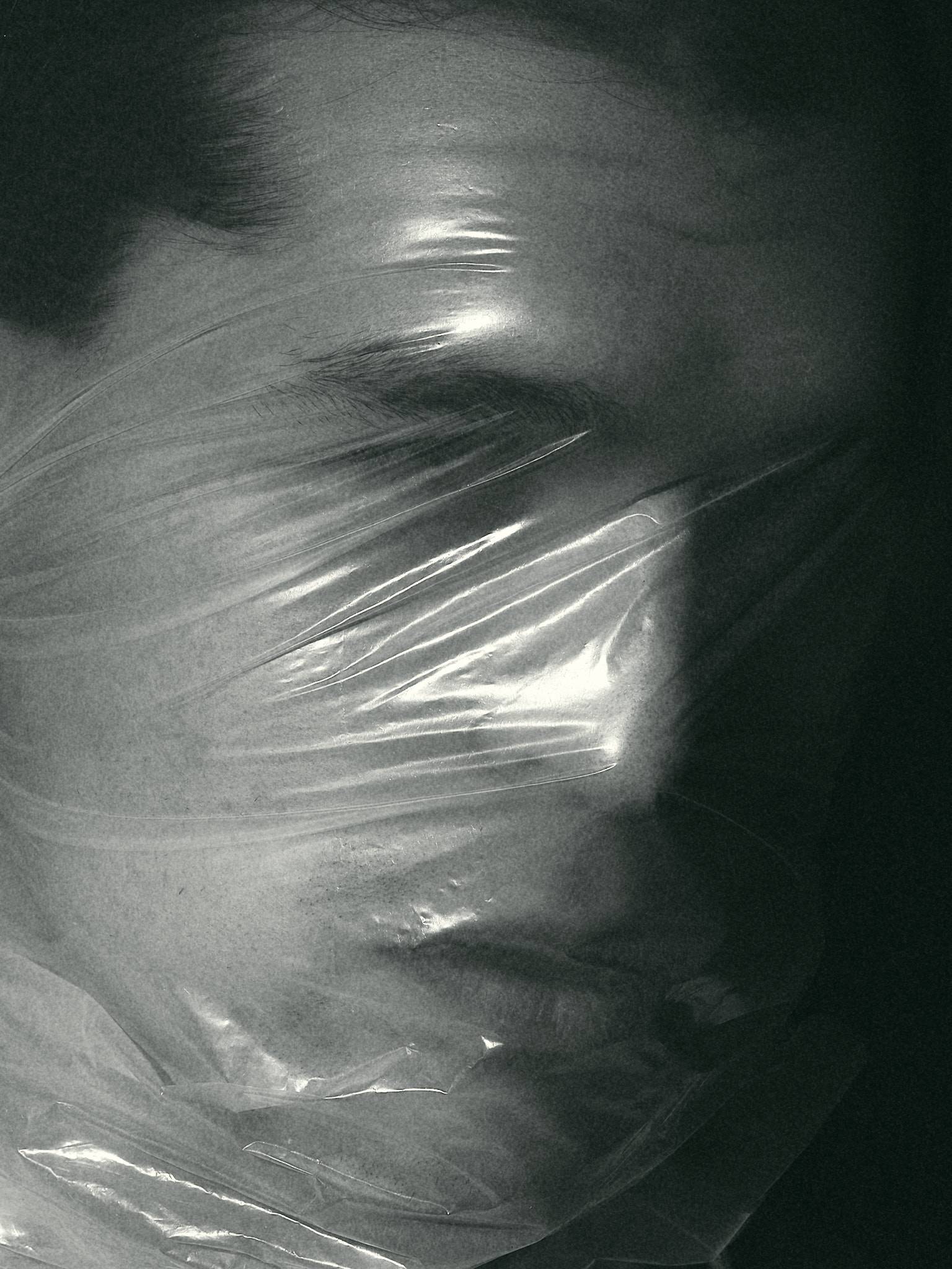 A face pressed beneath taut clear plastic, monochrome, the sheet catching light where it stretches over the features.