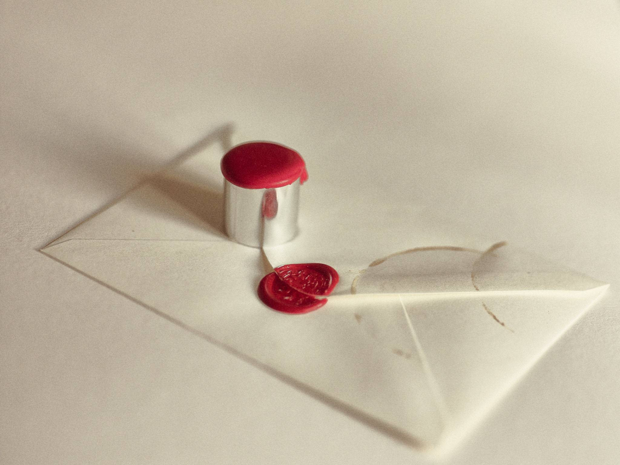 A silver sealing stamp stands upright on a cream envelope with red wax pooled over its face, a cracked wax seal beside it, lit high and soft.