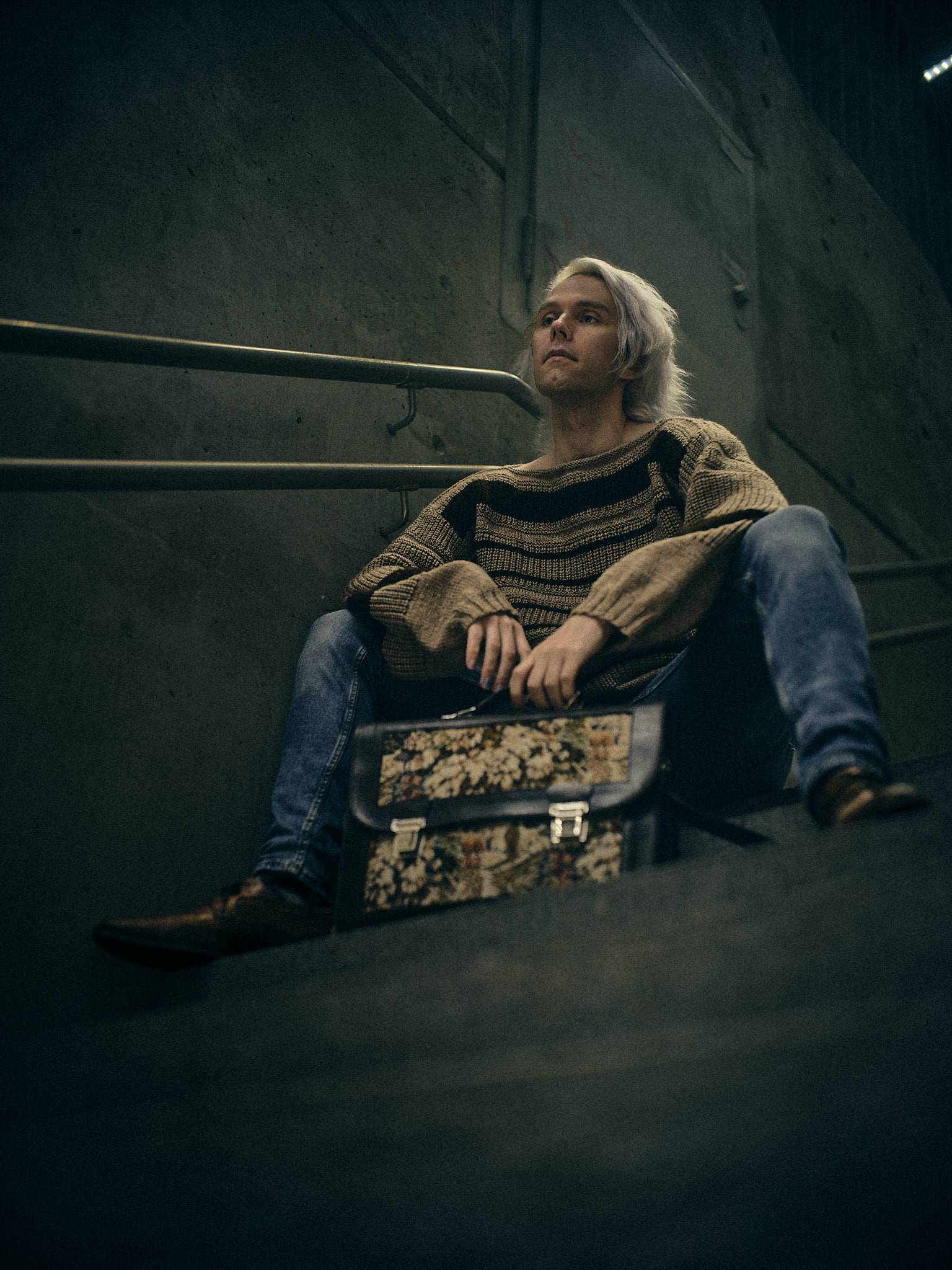 A figure seated low on concrete steps and leaning back, a floral tapestry briefcase propped against one leg, shot from below with a handrail crossing the wall behind.