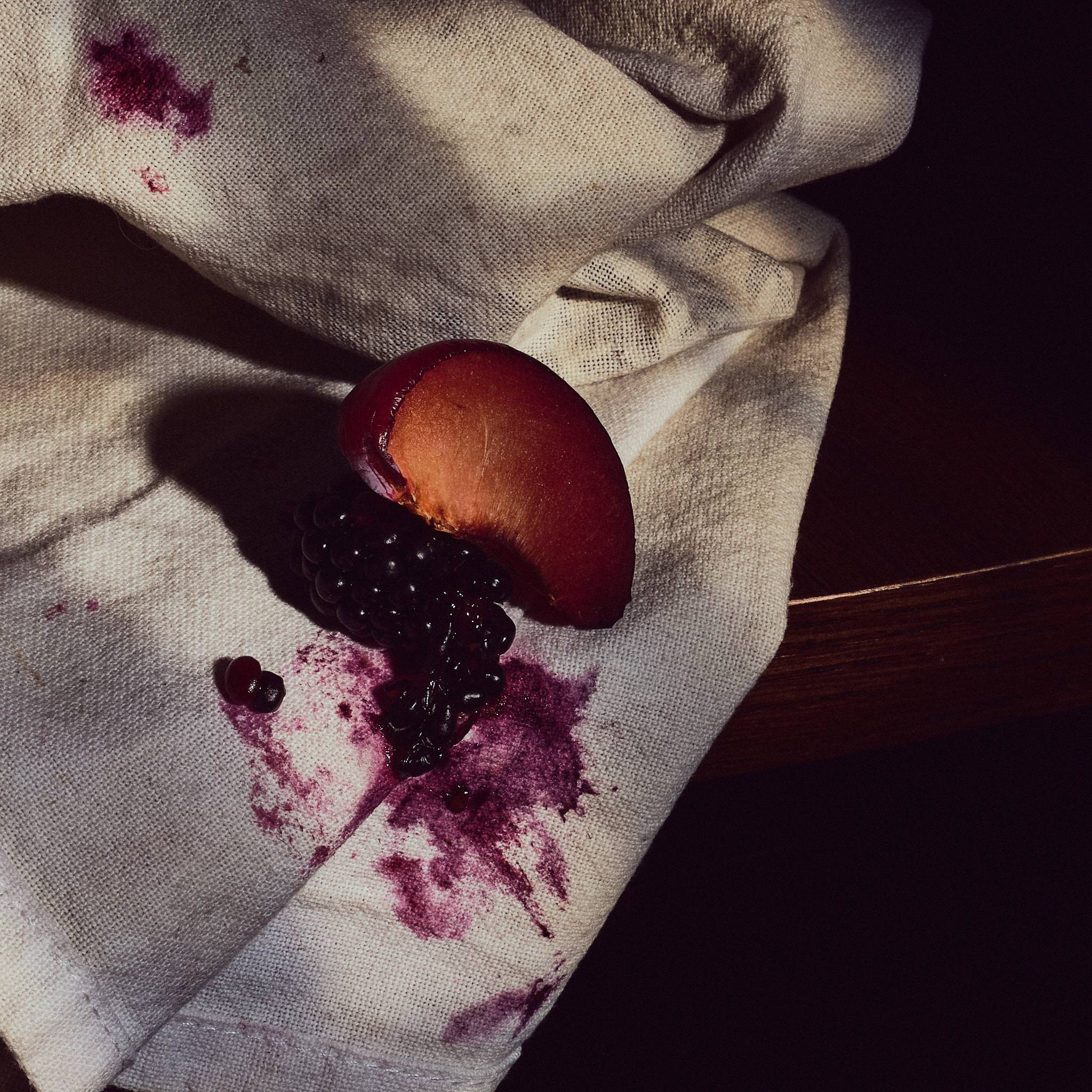 A wedge of plum and a crushed blackberry on rumpled cream linen, purple juice bled into the weave around them, lit hard from the left.