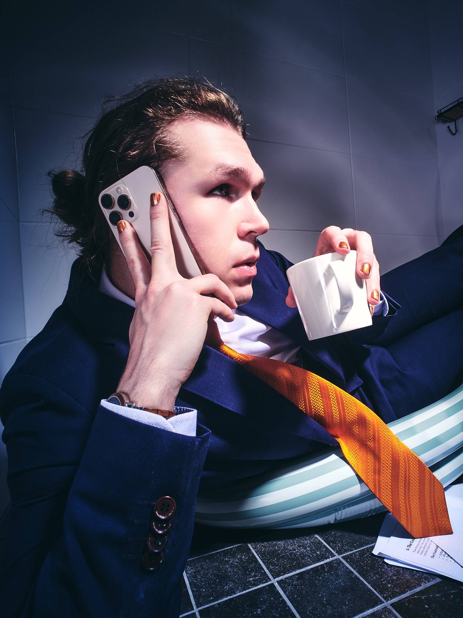 A man in a navy suit and orange tie sits in an inflatable paddling pool in a tiled bathroom, a phone to his ear and a coffee cup in his other hand.