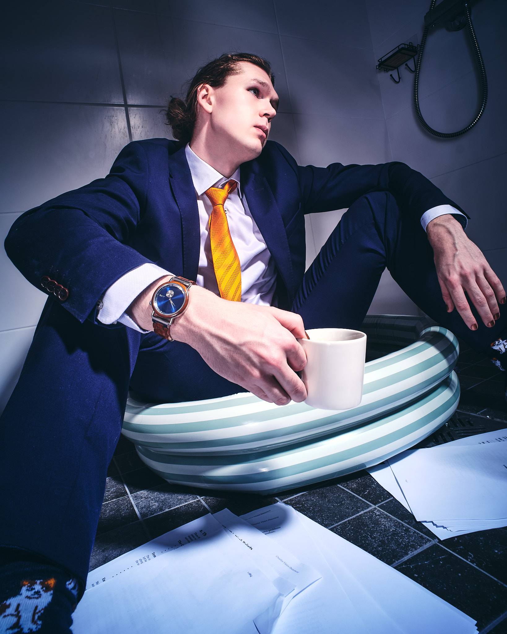 A man in a navy suit sits on a tiled bathroom floor, half inside an inflatable paddling pool, holding a coffee cup with documents spread around him.