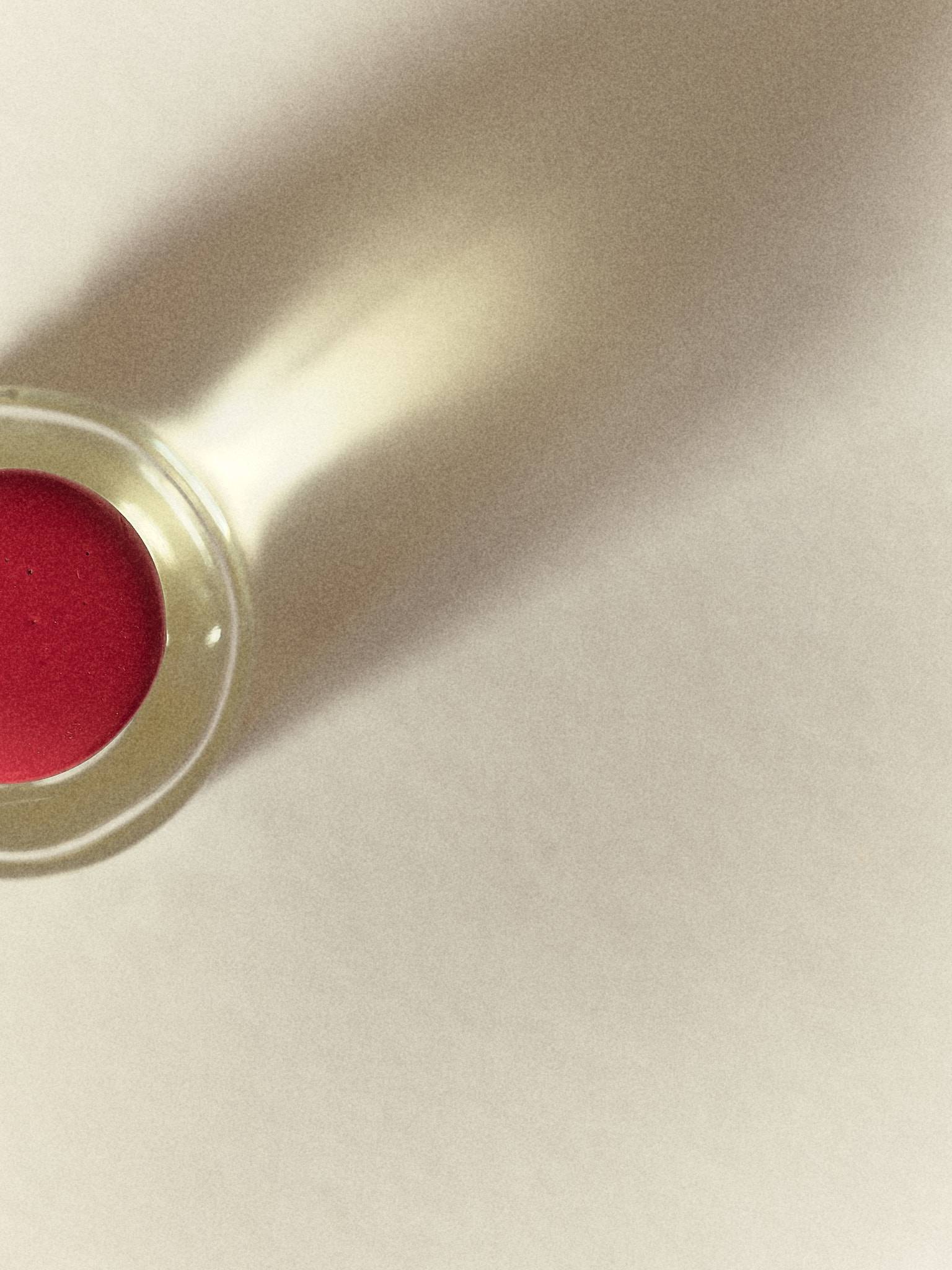 Looking down on a glass perfume bottle sealed with a disc of red wax, its long soft shadow thrown diagonally across a pale ground.