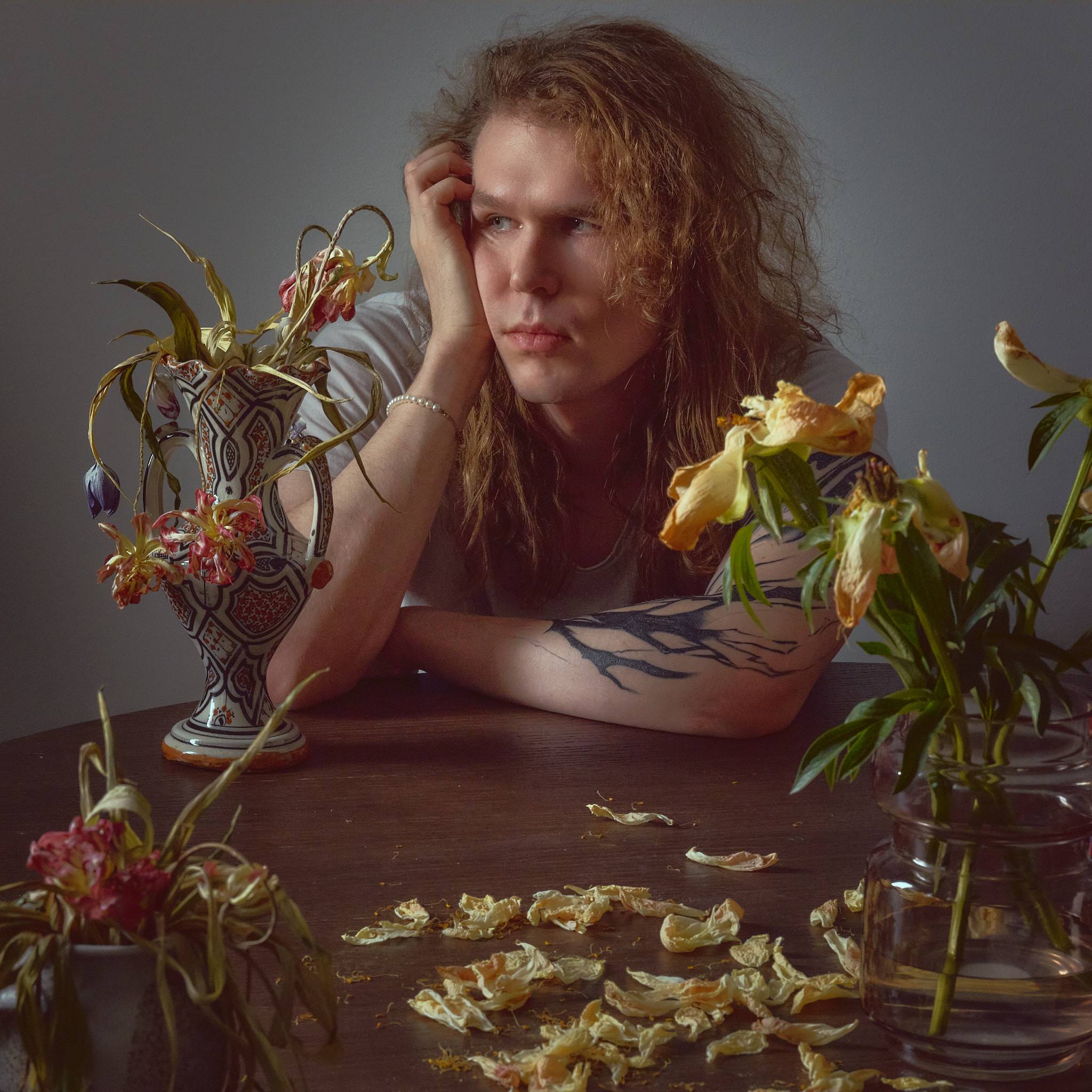 A person leans on a table surrounded by vases of collapsing lilies and tulips, dried petals scattered across the wood in front of them.