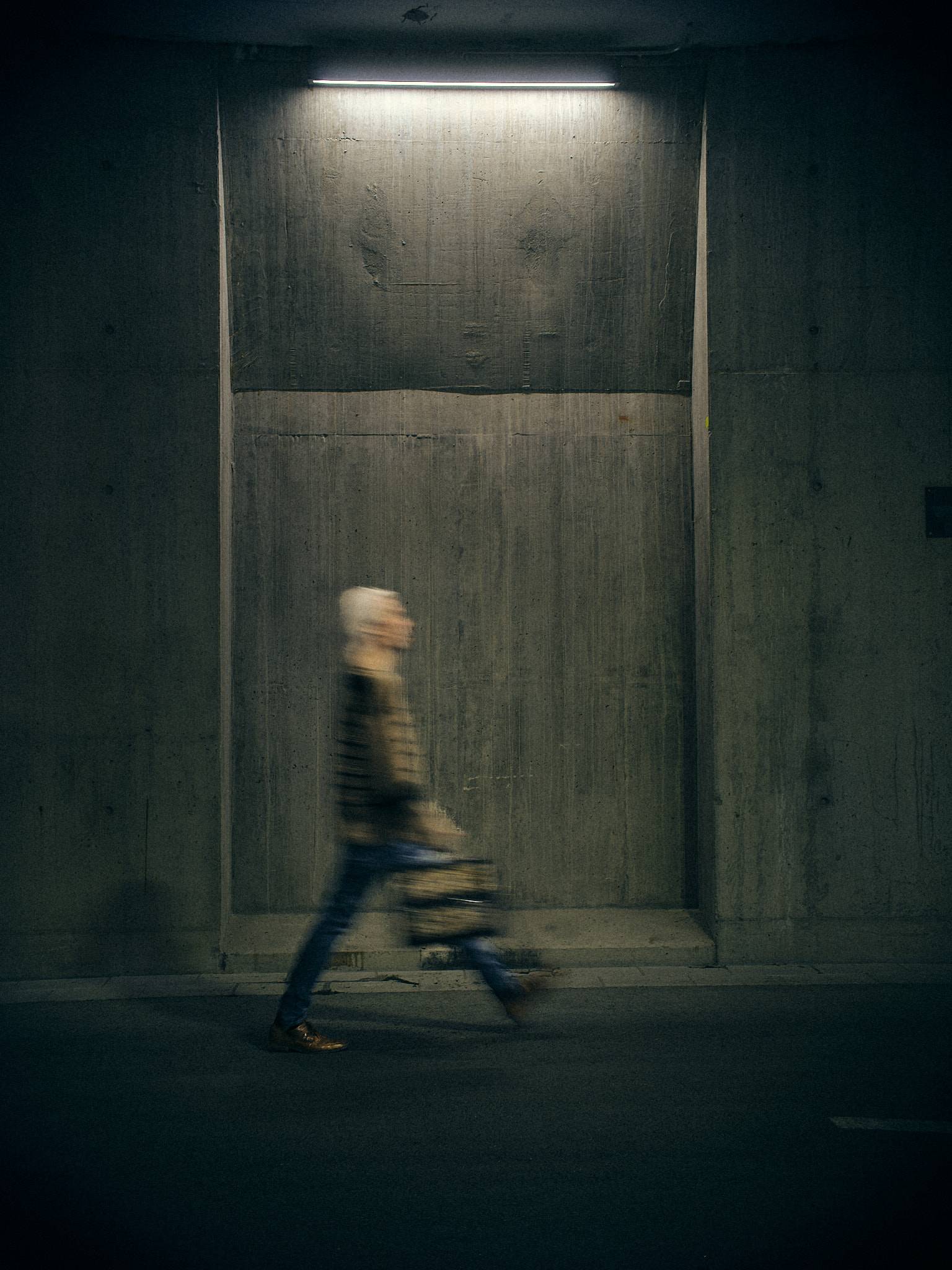 A figure walks through a lit concrete bay in an underpass, smeared by a long exposure, a single strip light burning above them.