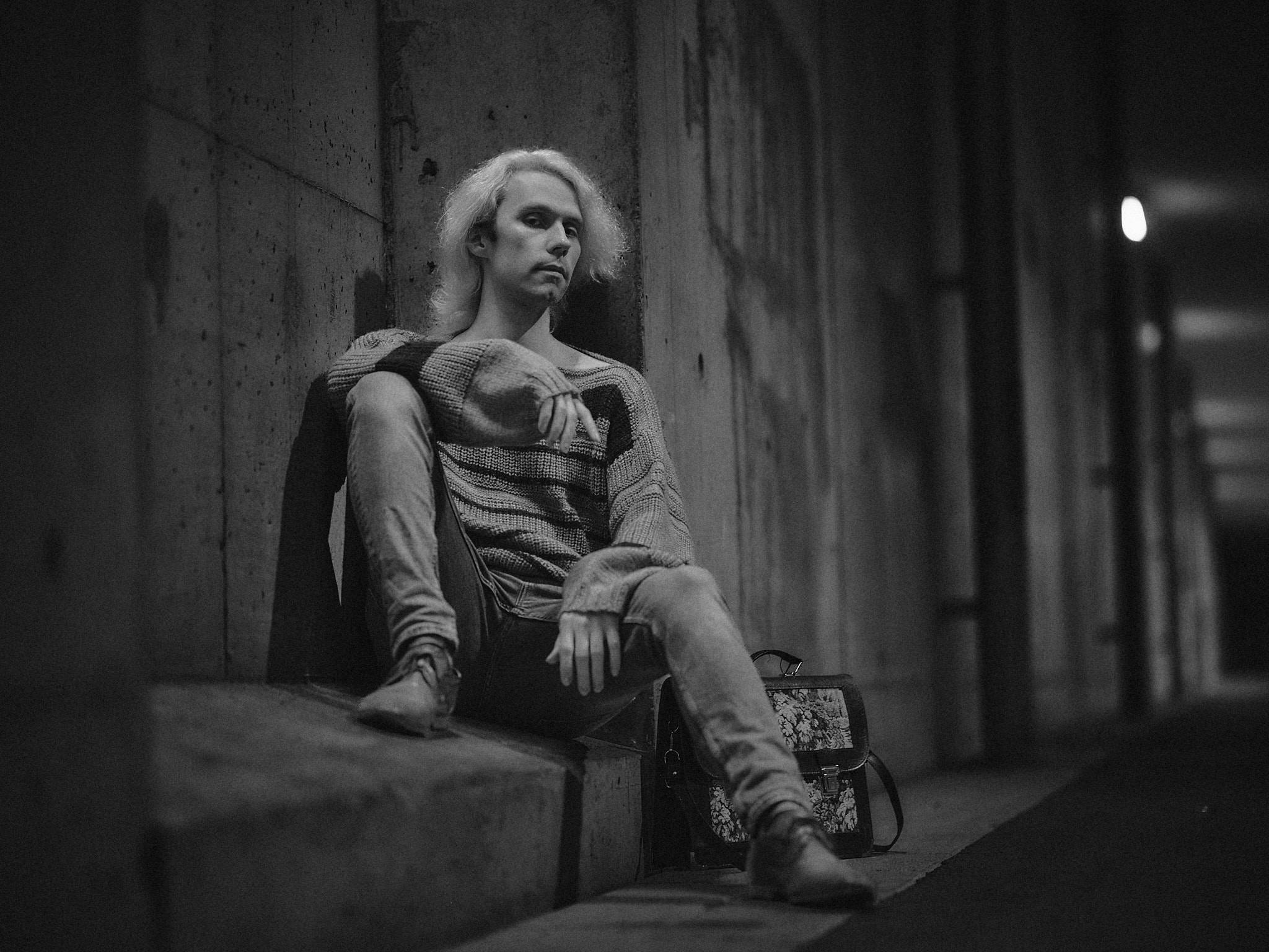 Monochrome. A figure seated on a concrete ledge in an underpass with a floral tapestry briefcase alongside, a lamp burning far down the tunnel behind.