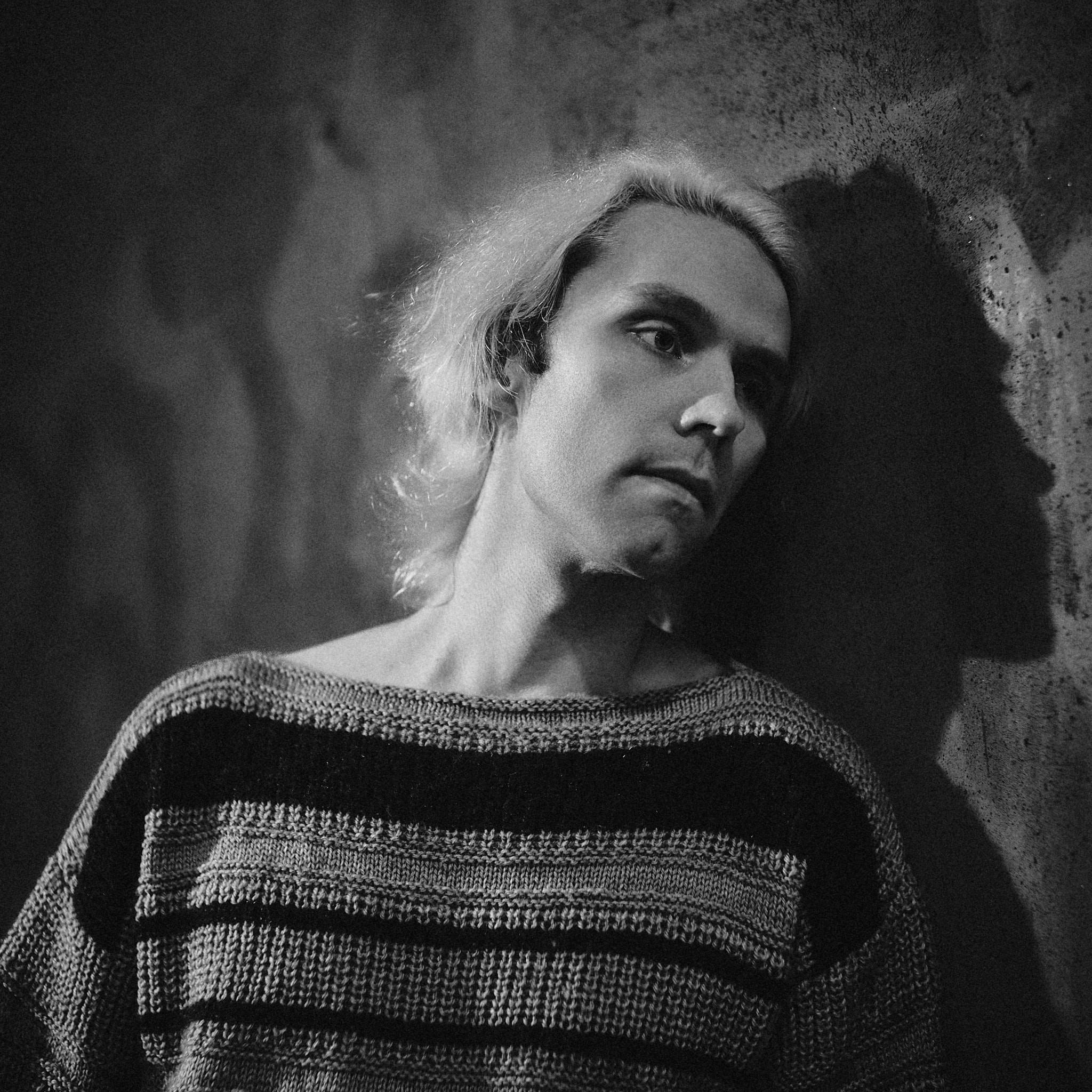Monochrome portrait against textured concrete, the sitter in a heavy striped knit, their own hard shadow doubling their profile on the wall behind.