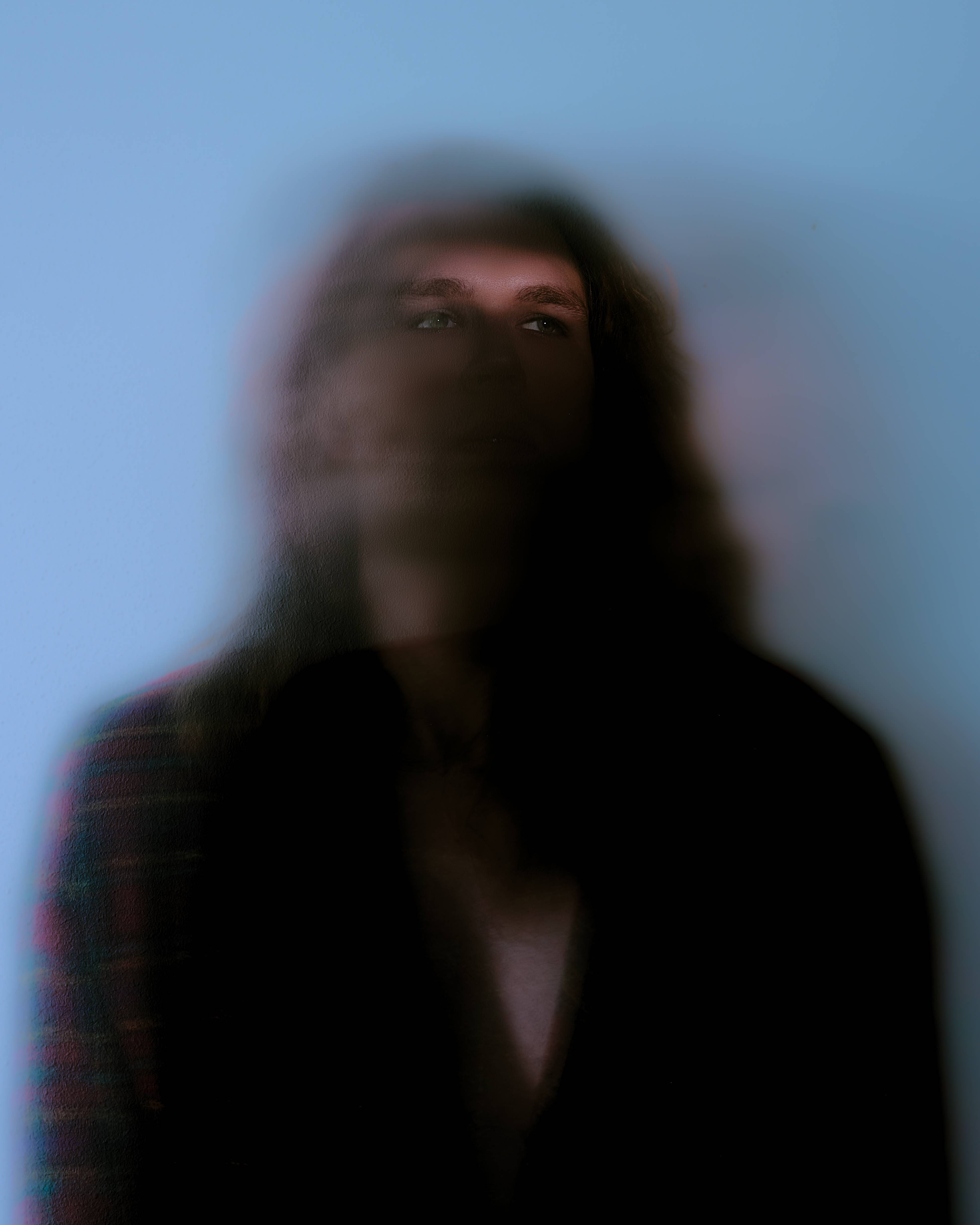 A long exposure of a figure turning, the features dissolved into movement against a pale blue wall.