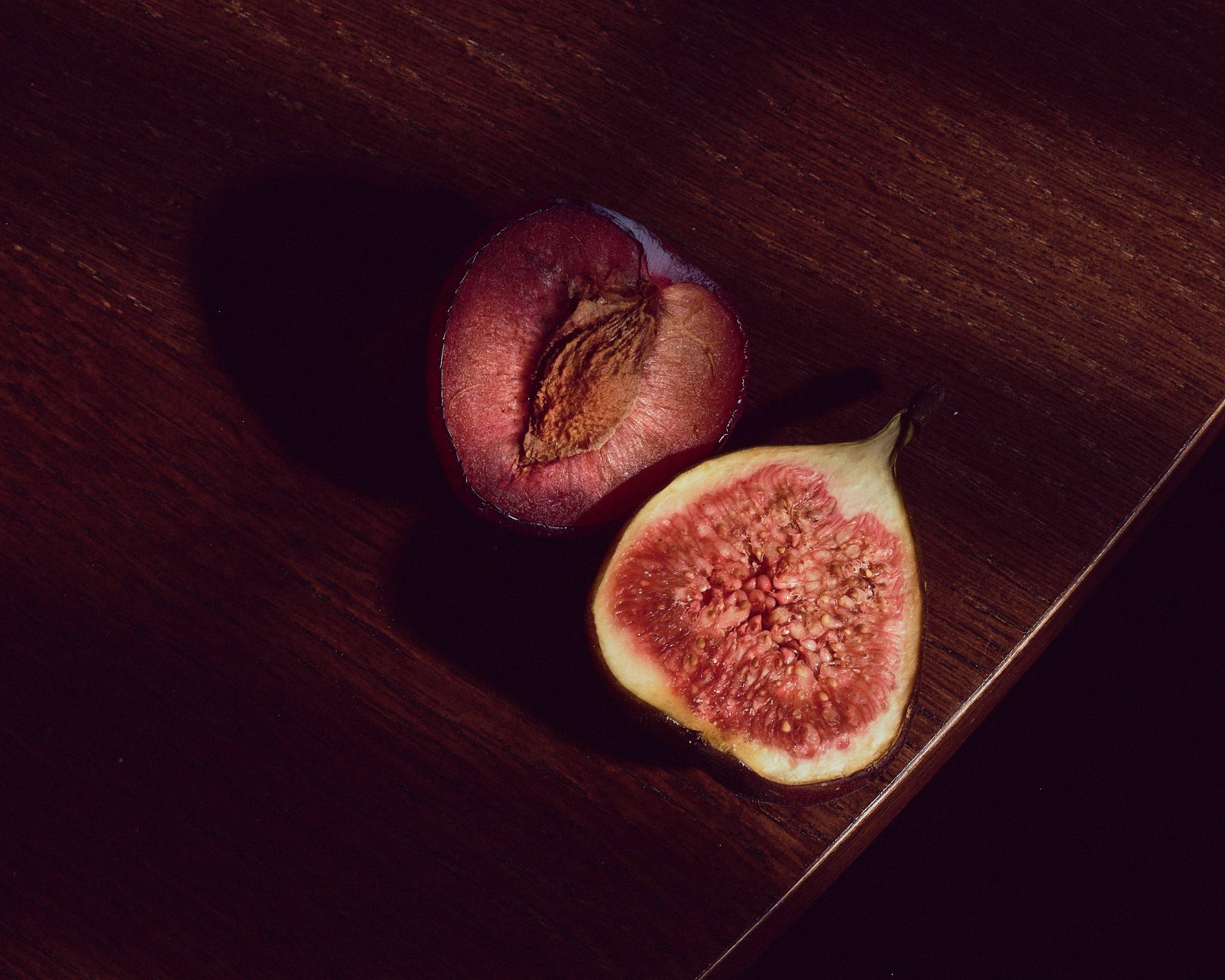 A halved plum and a halved fig lie on dark wood in a single shaft of light, their cut faces turned up, the rest of the table in shadow.