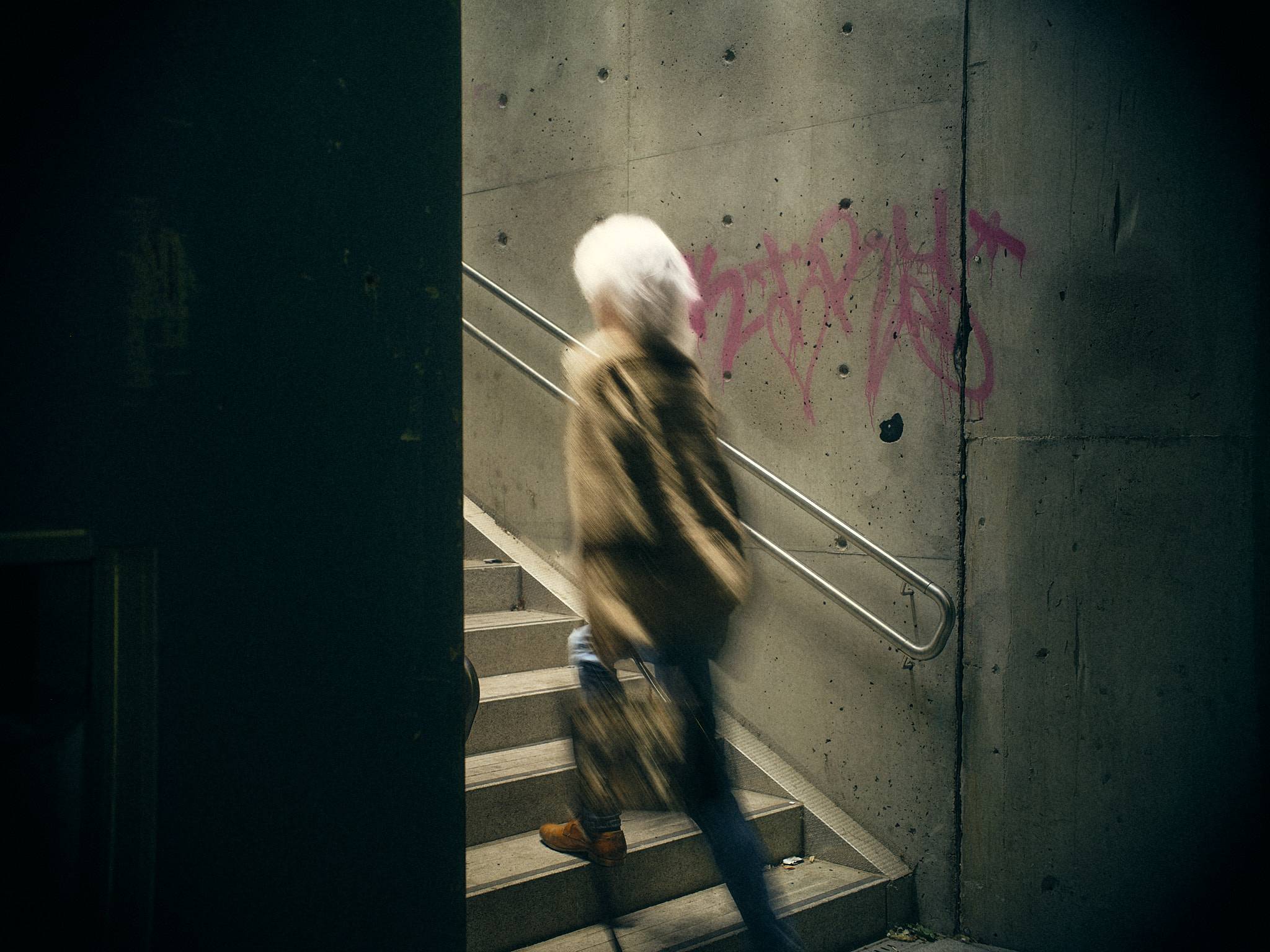 A figure climbs concrete stairs past pink graffiti, blurred by a long exposure, daylight falling down the stairwell from above.